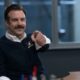 Jason Sudeikis como Ted Lasso sorrindo e apontando, vestindo suéter azul com brasão.