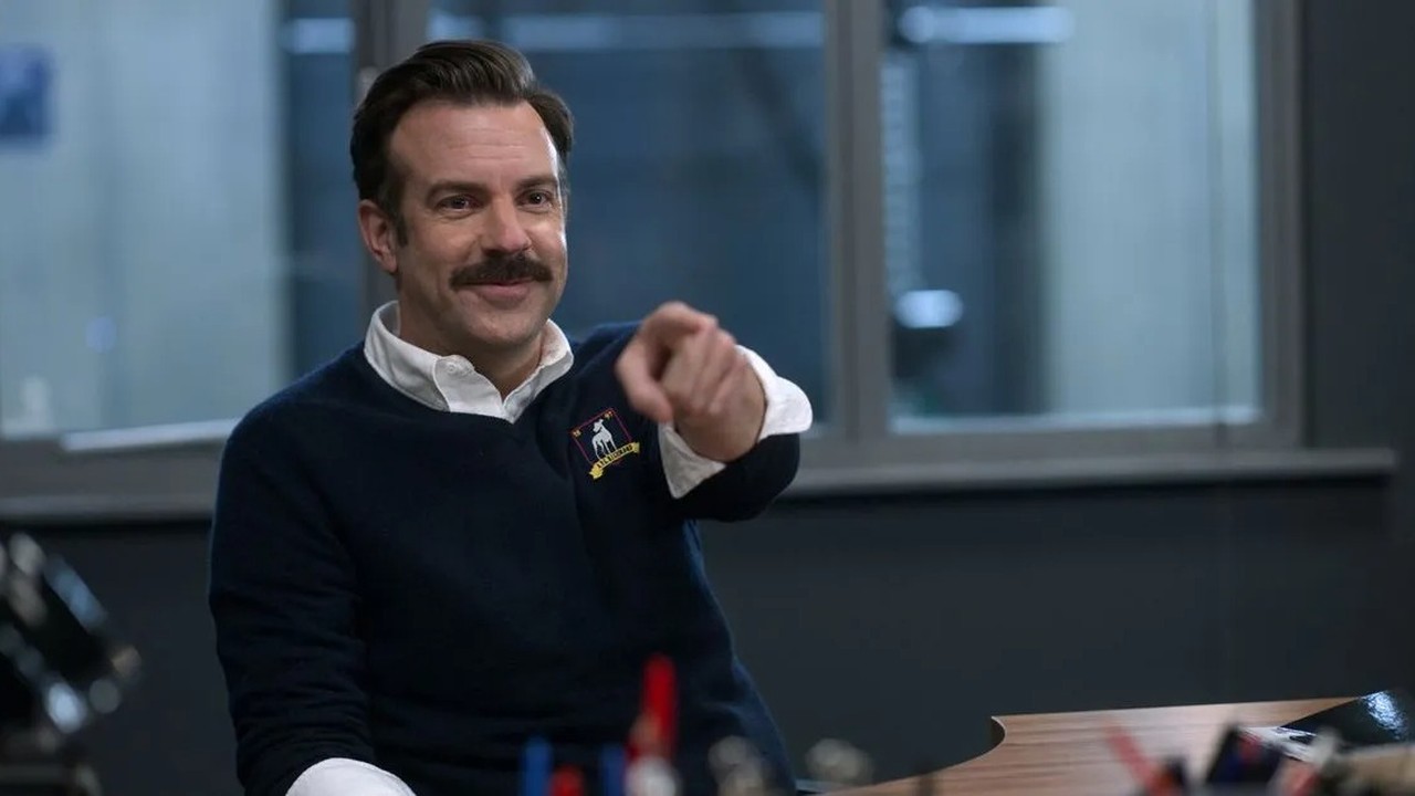 Jason Sudeikis como Ted Lasso sorrindo e apontando, vestindo suéter azul com brasão.