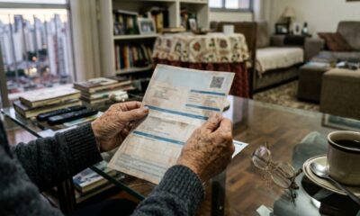 Mãos envelhecidas seguram uma conta ou boleto com texto borrado, sobre uma mesa de vidro.