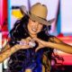 Cantora Ana Castela sorri, vestindo chapéu de cowboy, fazendo coração com as mãos no microfone em palco.