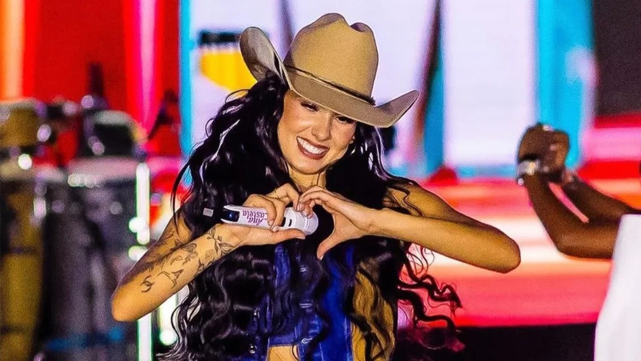 Cantora Ana Castela sorri, vestindo chapéu de cowboy, fazendo coração com as mãos no microfone em palco.