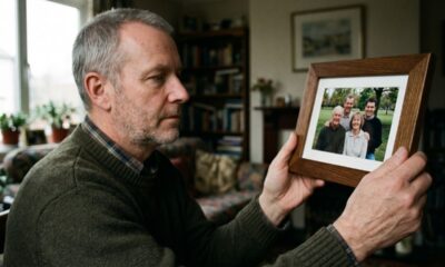 Homem com barba grisalha segura foto de família sorrindo, emoldurada, dentro de casa.