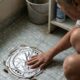 Mulher agachada cobrindo ralo do banheiro com papel alumínio, com baratas próximas.