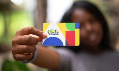 Mão segura cartão colorido do Programa Bolsa Família, com fundo desfocado.