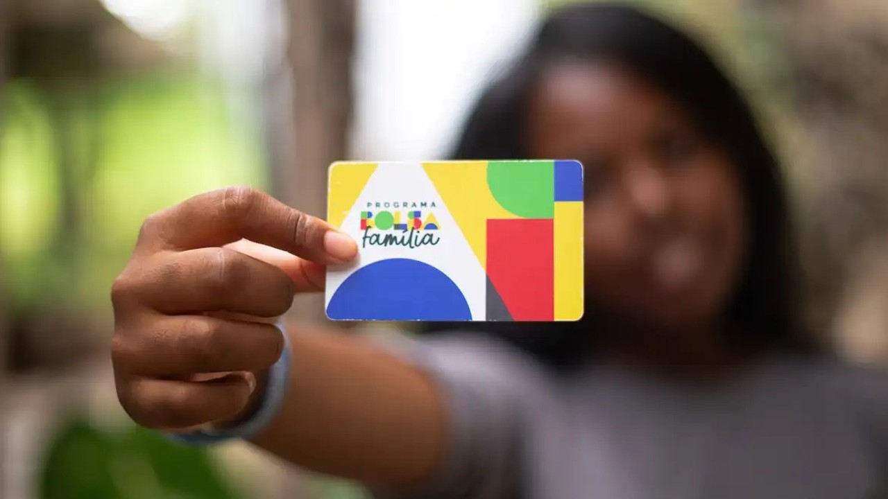 Mão segura cartão colorido do Programa Bolsa Família, com fundo desfocado.