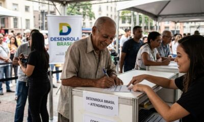 Homem idoso assina documento em balcão "Desenrola Brasil - Atendimento", assistido por mulher.