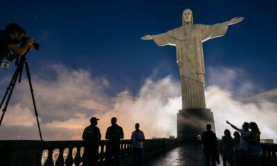 Cristo Redentor iluminado à noite, com fotógrafo e visitantes em primeiro plano.