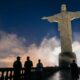 Cristo Redentor iluminado à noite, com fotógrafo e visitantes em primeiro plano.
