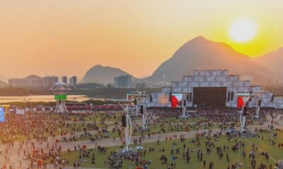 Festival de música ao pôr do sol. Palco principal, multidão, montanhas e rio ao fundo.