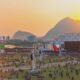 Festival de música ao pôr do sol. Palco principal, multidão, montanhas e rio ao fundo.