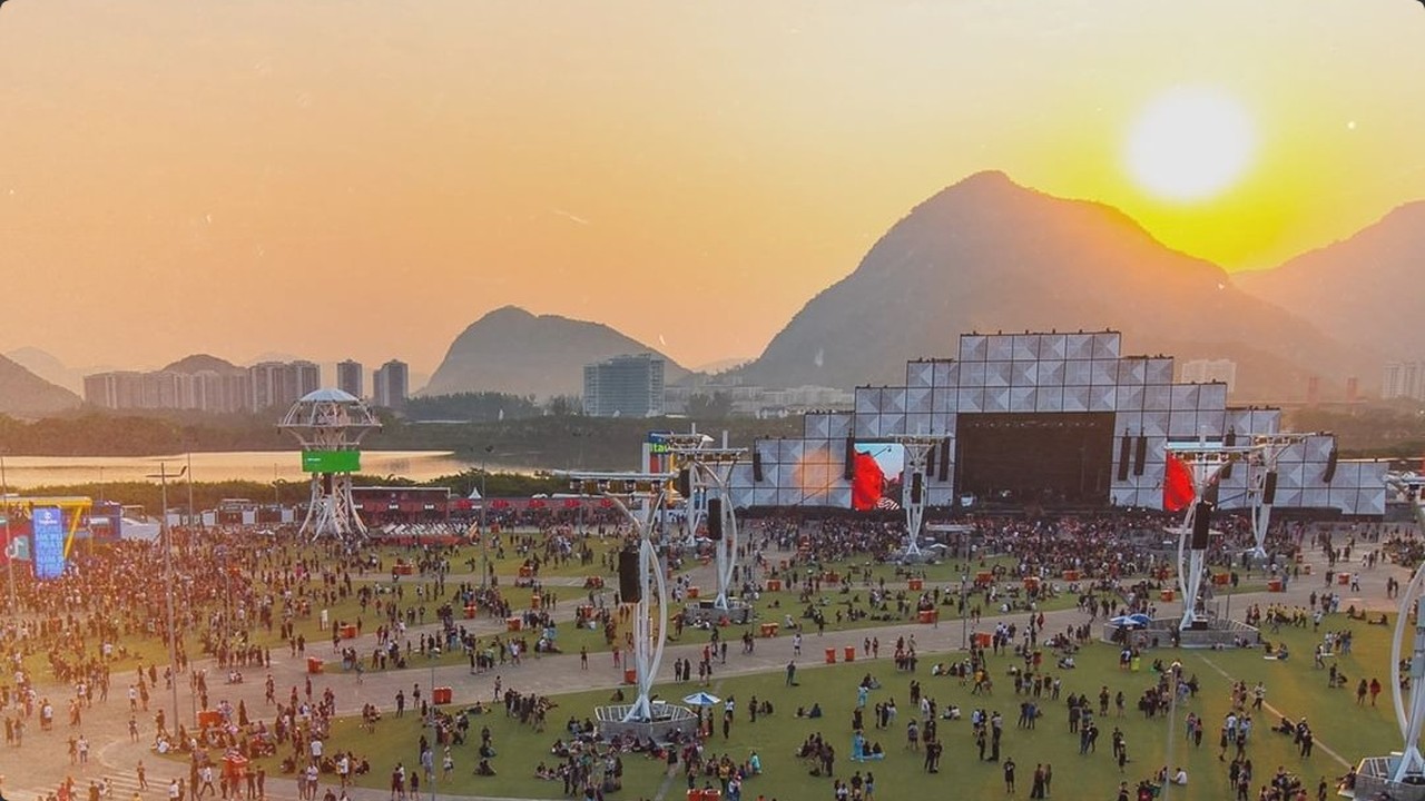 Festival de música ao pôr do sol. Palco principal, multidão, montanhas e rio ao fundo.