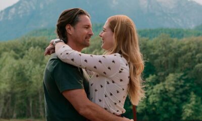 Homem e mulher sorrindo e abraçados, com montanhas verdes e floresta ao fundo.