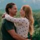 Homem e mulher sorrindo e abraçados, com montanhas verdes e floresta ao fundo.