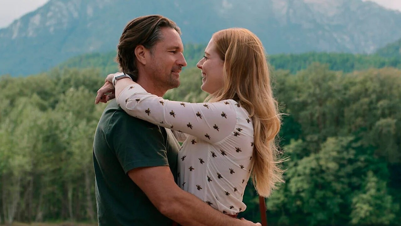 Homem e mulher sorrindo e abraçados, com montanhas verdes e floresta ao fundo.