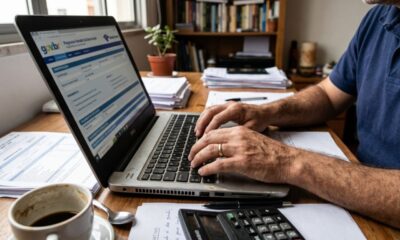 Pessoa digita em laptop com gov.br, "Programa Gerador da Declaração", calculadora e café na mesa.