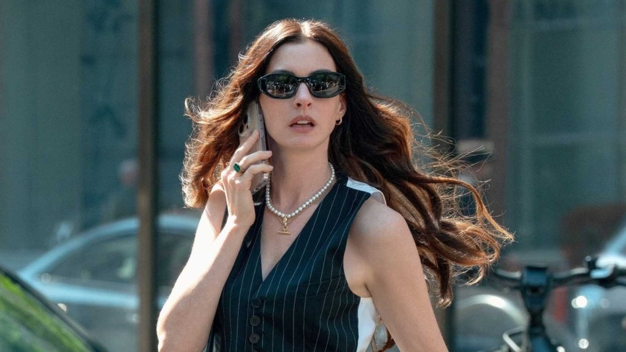 Anne Hathaway de óculos escuros e colete risca de giz, falando ao celular na rua.