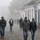 Várias pessoas agasalhadas caminham em rua de paralelepípedos sob forte neblina em cidade histórica.