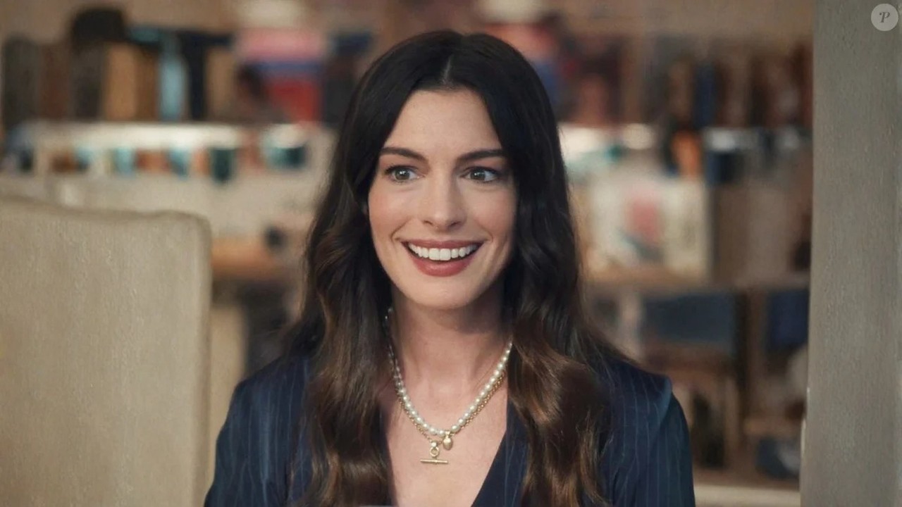 Close-up de Anne Hathaway sorrindo, com cabelo castanho escuro e colar de pérolas.