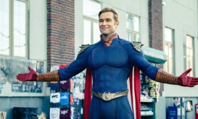 Homem sorridente em traje azul de super-herói com capa e luvas vermelhas, braços abertos.