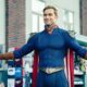 Homem sorridente em traje azul de super-herói com capa e luvas vermelhas, braços abertos.