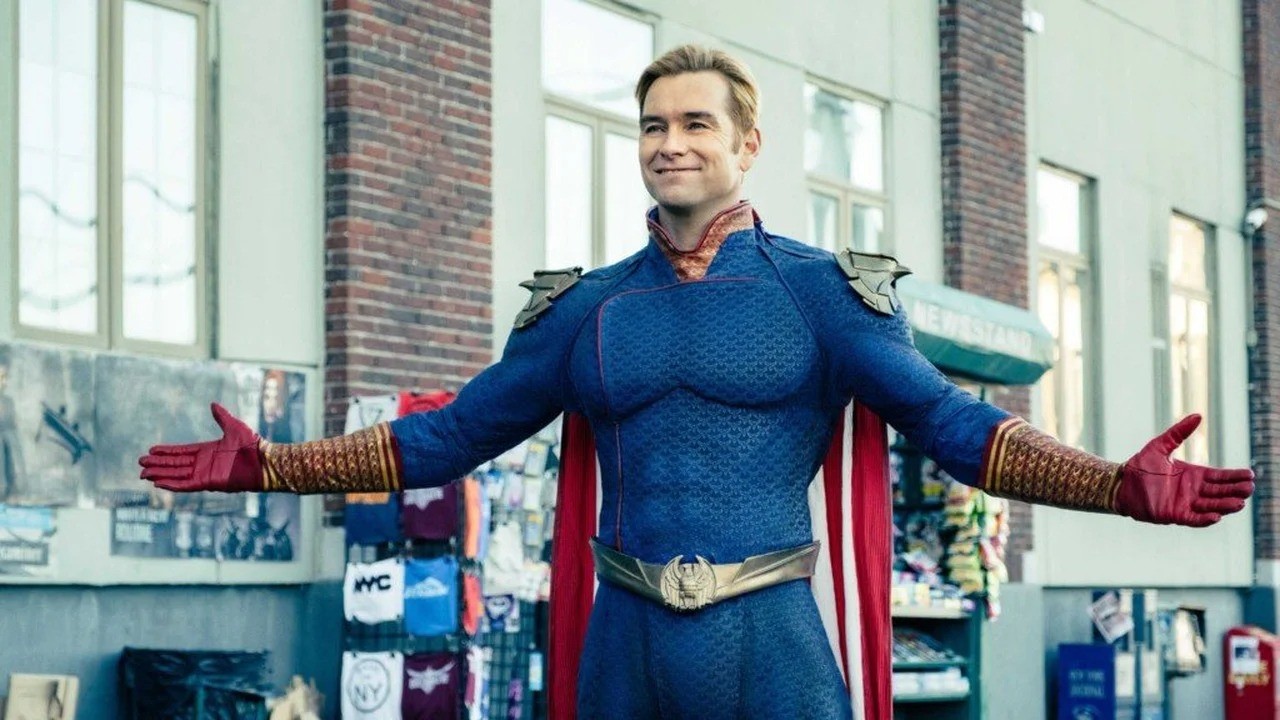 Homem sorridente em traje azul de super-herói com capa e luvas vermelhas, braços abertos.