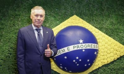 Carlo Ancelotti sorri, polegar para cima, ao lado do globo da bandeira brasileira.