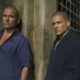 Dominic Purcell e Wentworth Miller, com cabeças raspadas, encostados em parede rústica com olhar sério.