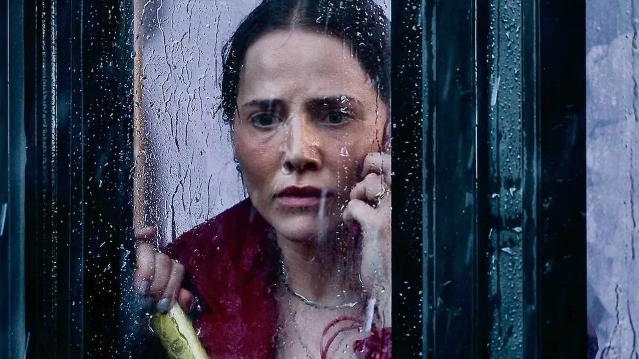 Letícia Colin (Adriana) com expressão angustiada olhando através de janela molhada pela chuva.