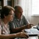 Dois idosos em casa usando um laptop. O homem aponta para a tela e a mulher usa o mouse.