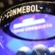 Recipiente de vidro com 'CONMEBOL' escrito e bolas azuis de sorteio 'Sudamericana'.