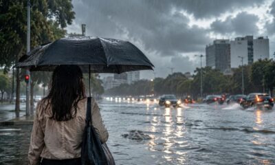 Pessoa com guarda-chuva em rua alagada com tráfego sob chuva forte e céu nublado.