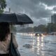 Pessoa com guarda-chuva em rua alagada com tráfego sob chuva forte e céu nublado.