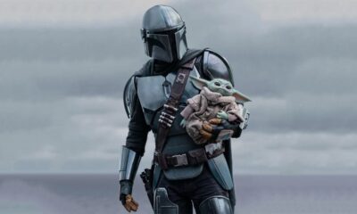 Mandalorian em armadura prateada segura Grogu (Baby Yoda). Céu nublado ao fundo.