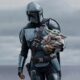 Mandalorian em armadura prateada segura Grogu (Baby Yoda). Céu nublado ao fundo.