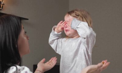 Criança loira chorando e cobrindo os olhos; adulto estende a mão com objeto.