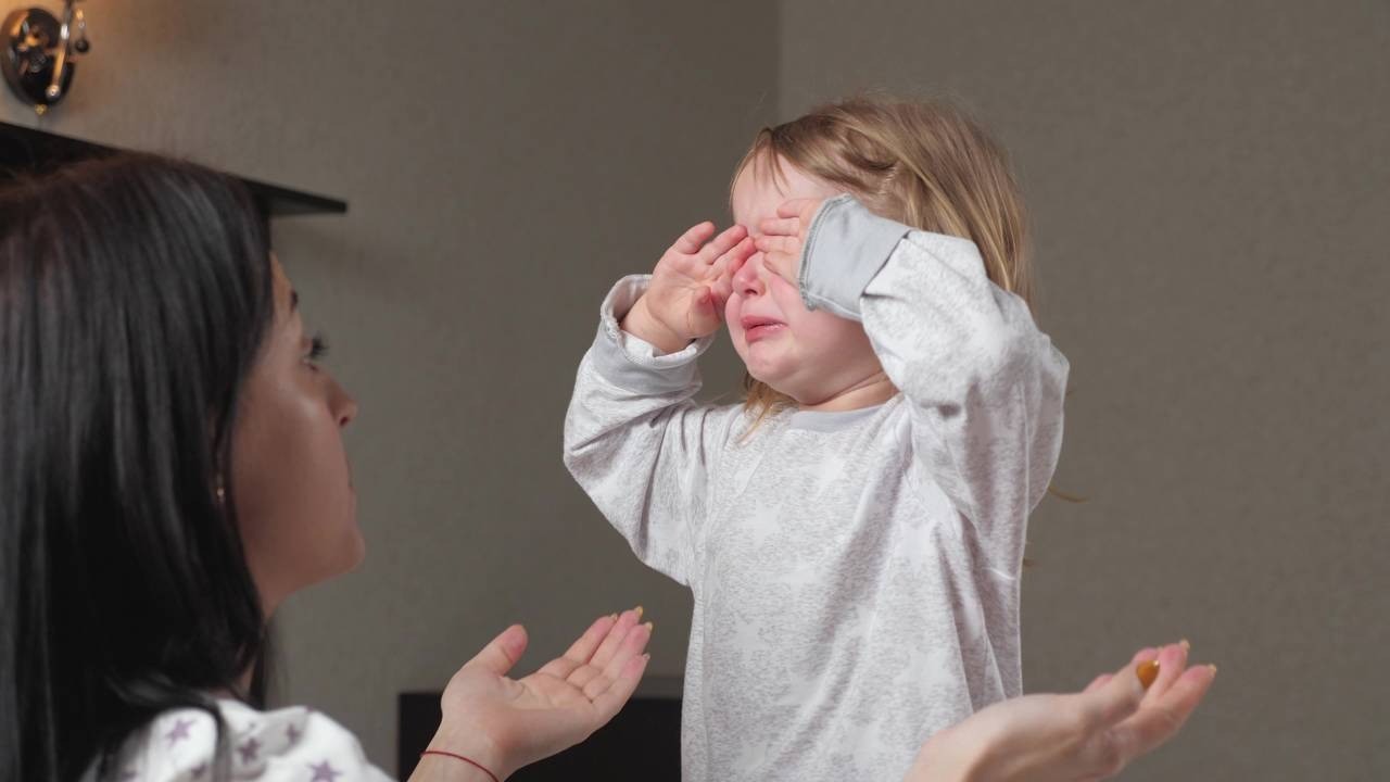 Criança loira chorando e cobrindo os olhos; adulto estende a mão com objeto.