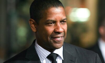 Denzel Washington sorrindo, usando terno escuro, camisa listrada e gravata preta.