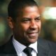Denzel Washington sorrindo, usando terno escuro, camisa listrada e gravata preta.