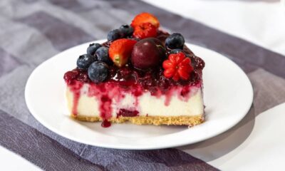 Fatia de cheesecake com calda de frutas vermelhas, mirtilos, morangos e uma pitanga.
