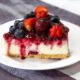 Fatia de cheesecake com calda de frutas vermelhas, mirtilos, morangos e uma pitanga.