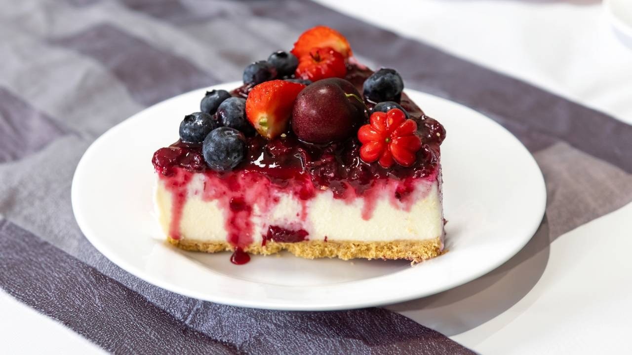 Fatia de cheesecake com calda de frutas vermelhas, mirtilos, morangos e uma pitanga.