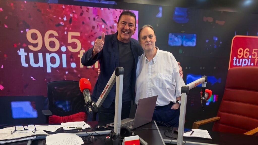 Mário Belisário e Heleno Rotay posam abraçados no estúdio da Super Rádio Tupi, com painel 96.5 tupi.fm ao fundo