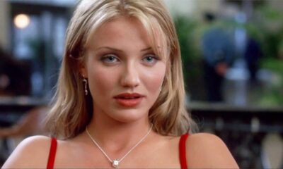 Cameron Diaz com cabelo loiro, olhos azuis, batom vermelho e colar prateado, fundo desfocado.
