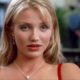 Cameron Diaz com cabelo loiro, olhos azuis, batom vermelho e colar prateado, fundo desfocado.