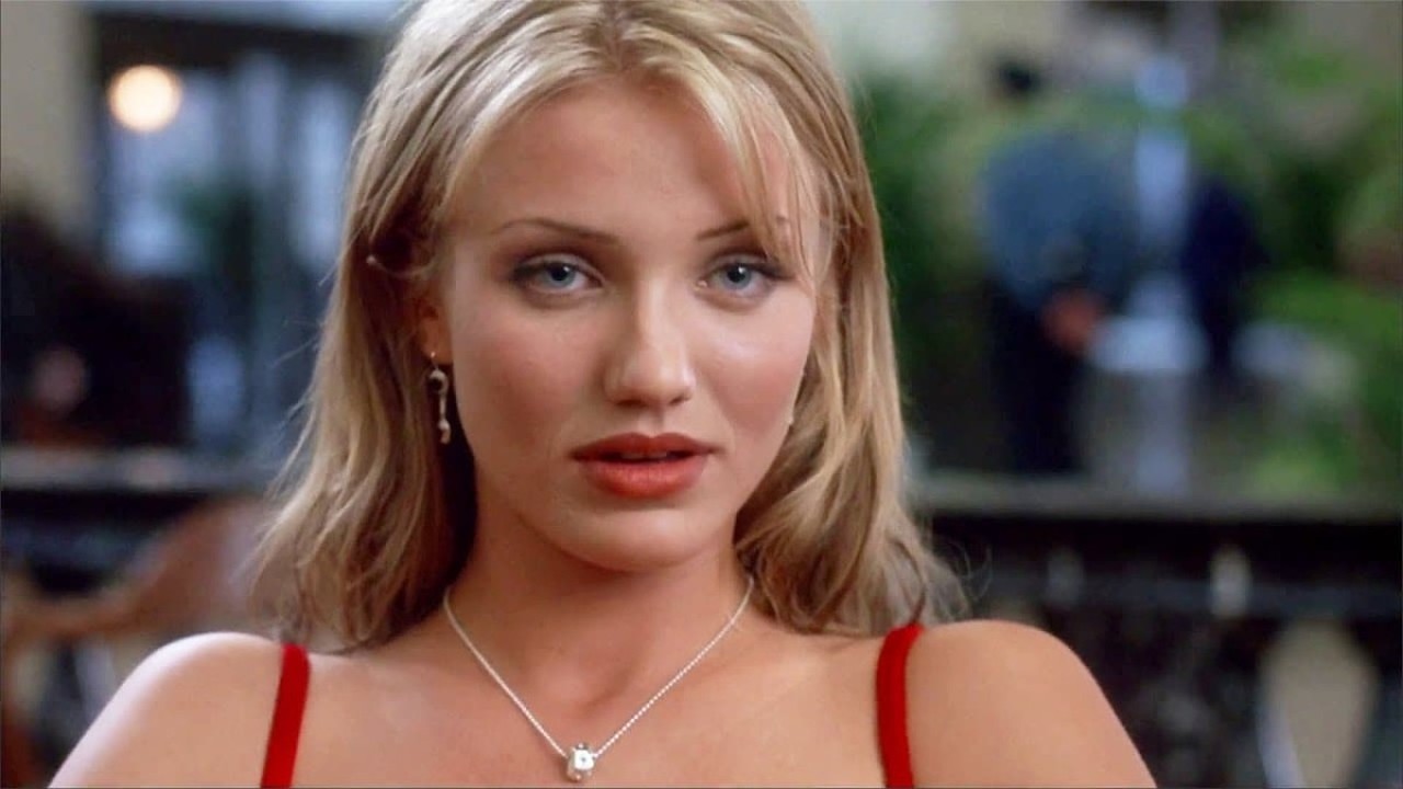 Cameron Diaz com cabelo loiro, olhos azuis, batom vermelho e colar prateado, fundo desfocado.