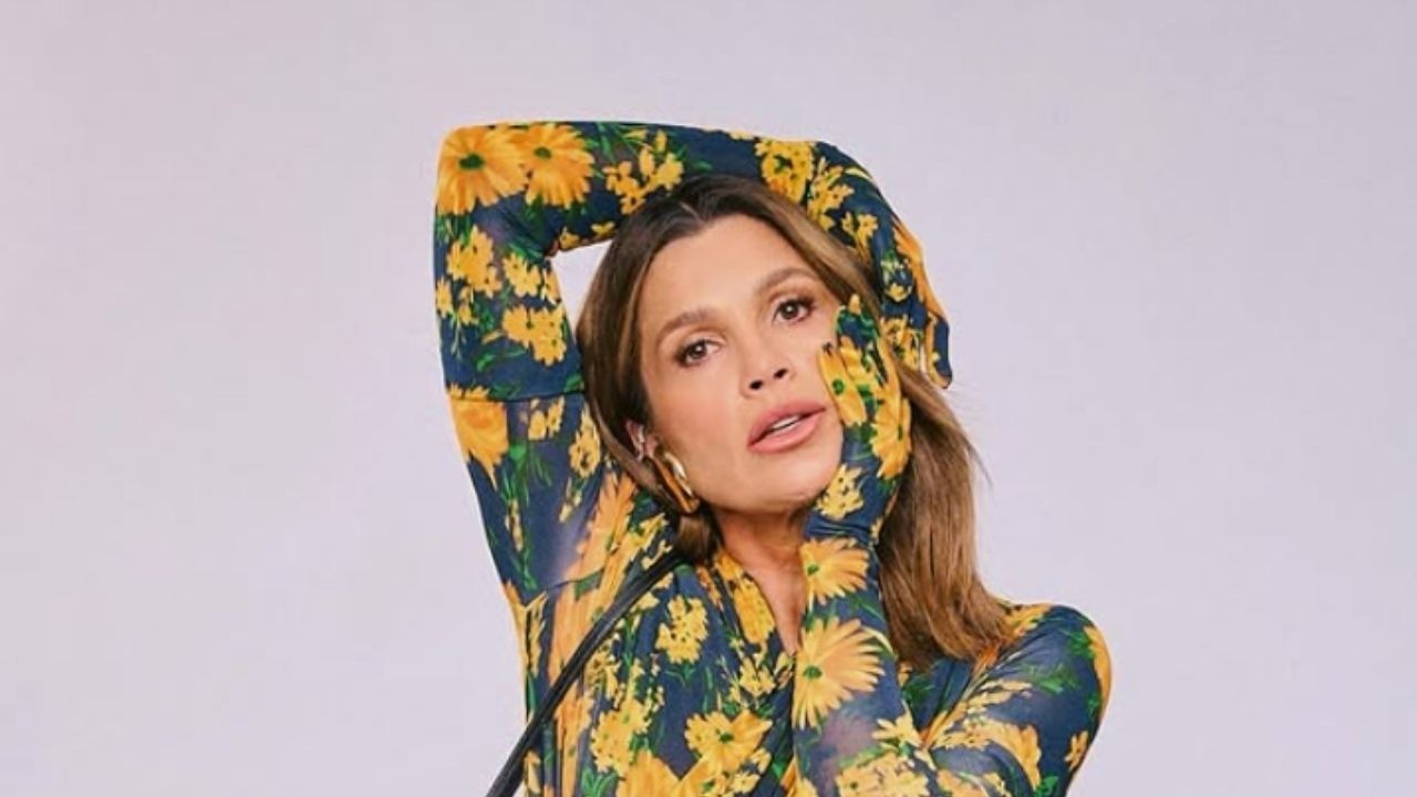 Atriz Flávia Alessandra em blusa florida azul e amarela, mãos no rosto, fundo claro.