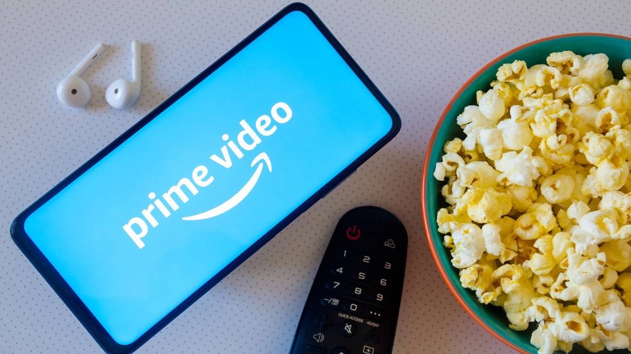 Celular com logo Prime Video, fones de ouvido, controle remoto e tigela de pipoca.
