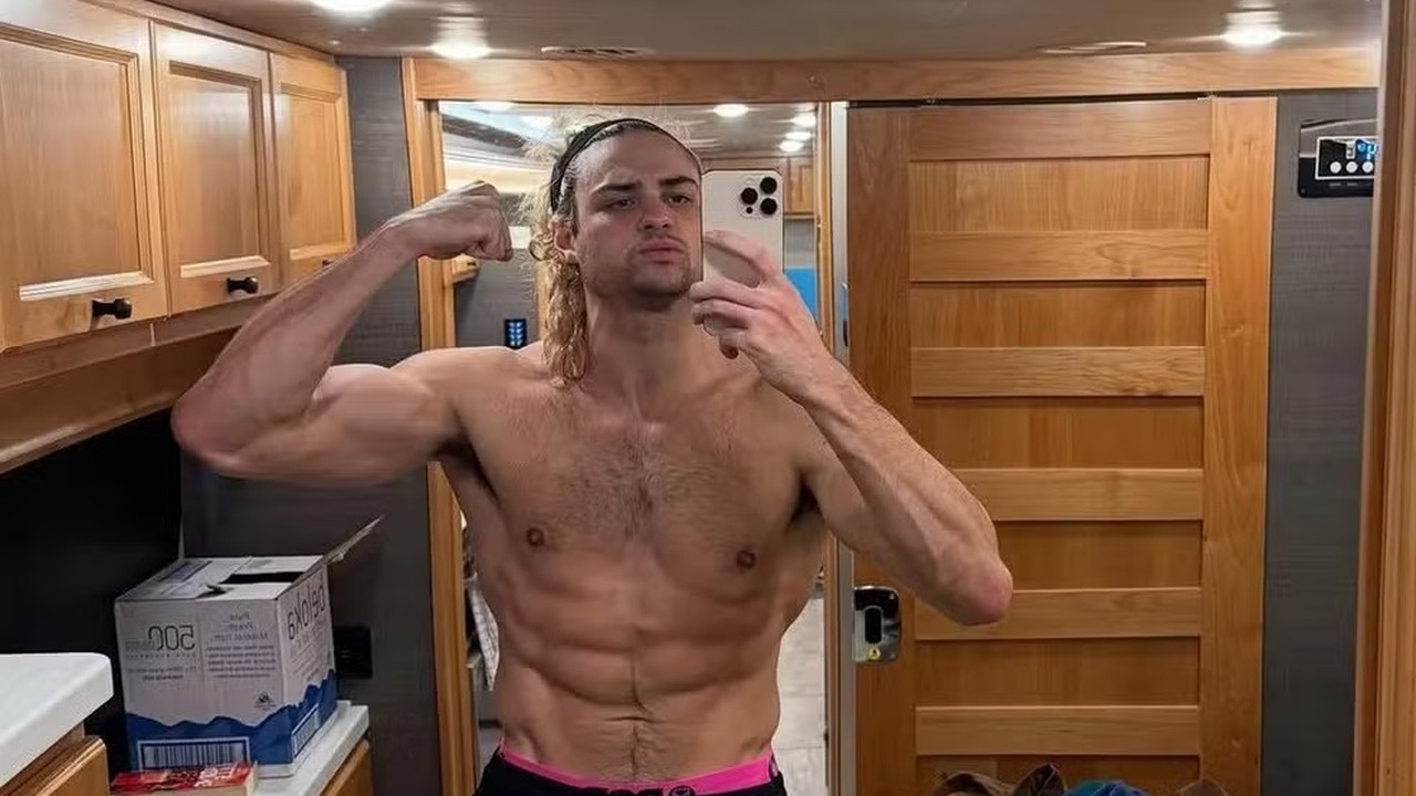 Noah Centineo sem camisa flexiona bíceps e abdômen em selfie no espelho de um trailer.