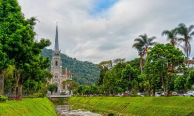 Catedral cinza gótica com torre alta e cruz, cercada por árvores verdes, montanhas e rio à frente.
