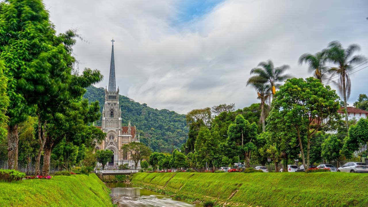 Catedral cinza gótica com torre alta e cruz, cercada por árvores verdes, montanhas e rio à frente.